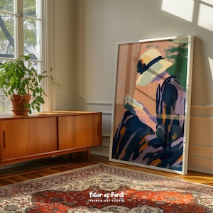 May include: A framed art print featuring a woman in a hat reading a book, rendered in soft pinks, blues, and greens. The artwork is leaning against a wall in a room with a wooden cabinet, a potted plant, and a patterned rug. The text "tiger of Paris FRENCH ART STUDIO" is visible.