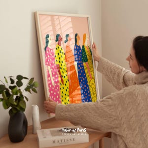May include: A framed art print featuring a colorful illustration of five women in polka-dot dresses. The dresses are in shades of pink, yellow, orange, blue, and green. The artwork is displayed in a white frame and is being held by a person.