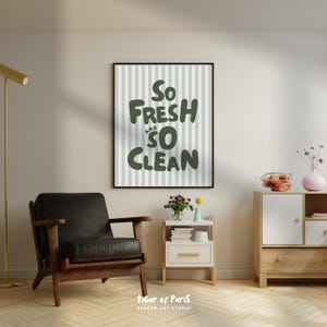May include: A framed wall art print with the words "So Fresh and So Clean" in dark green lettering. The background has vertical light gray and white stripes. A black leather armchair and a wooden cabinet are in the foreground.