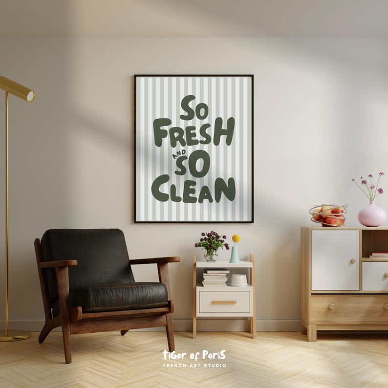 May include: A framed wall art print with the words "So Fresh and So Clean" in dark green lettering. The background has vertical light gray and white stripes. A black leather armchair and a wooden cabinet are in the foreground.