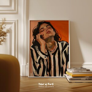 May include: A framed art print featuring a portrait of a person with dark hair and closed eyes, wearing a black and white striped shirt. The background is orange. The text "tiger of Paris FRENCH ART STUDIO" is at the bottom.