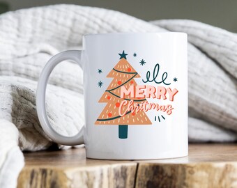 May include: White ceramic mug with a Christmas tree design and the text "Jingle Bell Merry Christmas". The mug is sitting on a wooden surface.