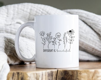 May include: White ceramic mug with a black outline of flowers and the text "Different is beautiful". The mug is sitting on a wooden coaster.