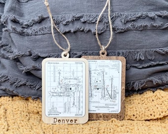 May include: Two wooden ornaments with aviation chart designs. One ornament reads "Denver" and the other reads "Lake City". Both have a natural twine loop for hanging. The background includes a gray pillow and a yellow blanket.