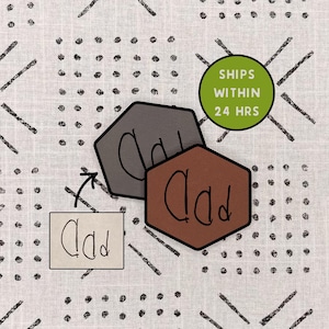 May include: Two hexagonal-shaped objects, one gray and one brown, with the word "Dad" written on them. A small white rectangle with the word "Dad" is also present. The background features a white fabric with a black geometric pattern.