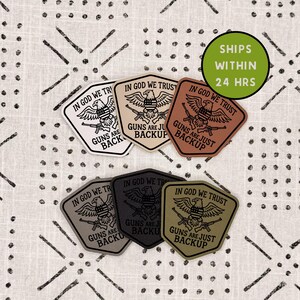 May include: A collection of six shield-shaped patches in white, tan, brown, gray, black, and olive green. Each patch features a graphic of an eagle with a shield, a rifle, and the text IN GOD WE TRUST GUNS ARE JUST BACKUP.