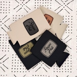 May include: Four foam beverage insulators in beige and black, each featuring a rectangular patch with the text Raisin Hell. The patches are in varying colors including dark gray, brown, olive green, and light gray, centered on the insulators.