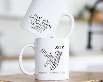 May include: Two white ceramic mugs stacked on a wooden surface. The top mug has text that reads "First Solo Marissa Swartz 10/24/2018 PA-28-161 N370TC." The bottom mug features a black and white airport diagram and the text "SGF Springfield-Branson Intl."