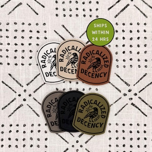 May include: Six shield-shaped patches in white, beige, brown, gray, black, and olive green. Each patch features a black graphic of a bird holding a branch, surrounded by the text "RADICALIZED BY DECENCY" and "BASIC" in a circular arrangement.