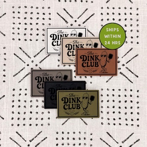 May include: A collection of six rectangular patches in various colors including white, tan, brown, gray, black, and olive green. Each patch features the text The DINK CLUB, a cartoon pickle wearing sunglasses and sneakers, and a pickleball paddle.
