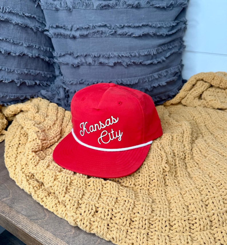 May include: A red baseball cap with the words "Kansas City" embroidered in white script. The cap has a white rope detail and a curved brim. The hat is resting on a yellow knitted blanket.