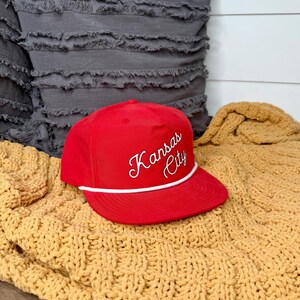 May include: A red baseball cap with the words "Kansas City" embroidered in white script. The cap has a white rope detail along the brim and is displayed on a yellow knitted blanket.