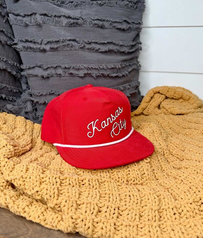 May include: A red baseball cap with the words "Kansas City" embroidered in white script. The cap has a white rope detail along the brim and is displayed on a yellow knitted blanket.