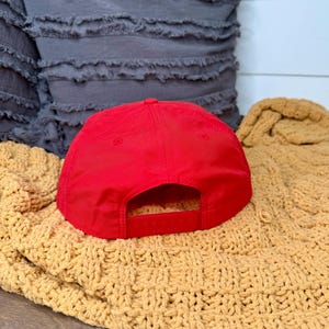 May include: A bright red baseball cap with a flat brim and adjustable snap closure. The cap is resting on a textured, mustard-colored blanket. A gray pillow is in the background.
