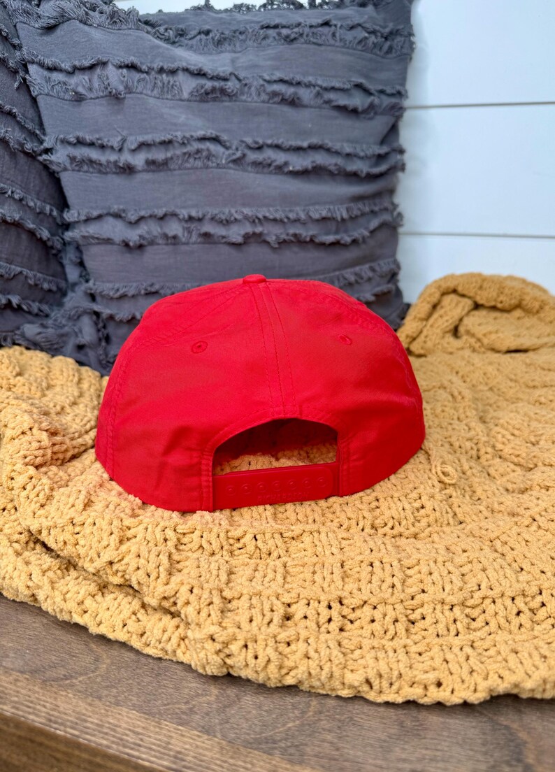 May include: A bright red baseball cap with a flat brim and adjustable snap closure. The cap is resting on a textured, mustard-colored blanket. A gray pillow is in the background.