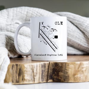 May include: White ceramic mug featuring a black and white airport diagram with the letters "CLE" and "Cleveland-Hopkins Intl" printed on it. The mug has a curved handle and is set on a wooden surface.