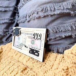 May include: A wooden frame with a graduation cap, the name "AVA" and the text "Class of 2025" in black lettering. The frame is designed to hold a dollar bill. The frame is sitting on a yellow knitted blanket.