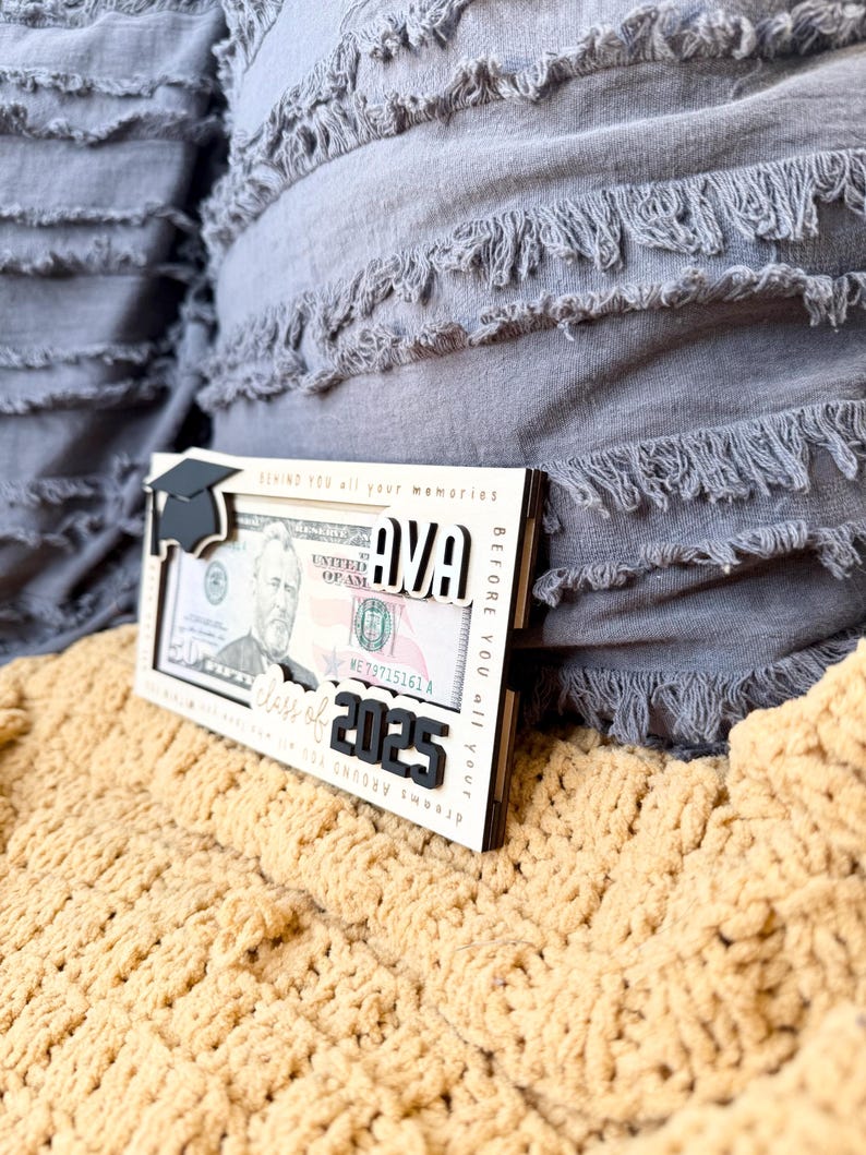 May include: A wooden frame with a graduation cap, the name "AVA" and the text "Class of 2025" in black lettering. The frame is designed to hold a dollar bill. The frame is sitting on a yellow knitted blanket.