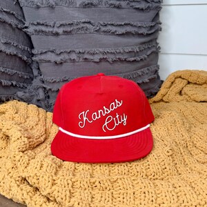 May include: A red baseball cap with the words "Kansas City" in white script. The cap has a white rope detail and a flat brim. The hat is displayed on a yellow knitted blanket.
