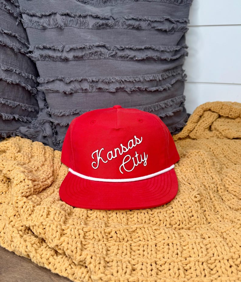 May include: A red baseball cap with the words "Kansas City" in white script. The cap has a white rope detail and a flat brim. The hat is displayed on a yellow knitted blanket.