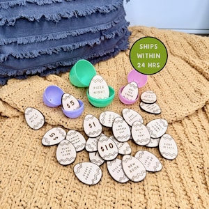 May include: Wooden egg-shaped tokens with various activities and dollar amounts, scattered on a tan knitted blanket. The tokens include phrases like "Pizza Night," "Family Movie Night," and "$10." Colorful plastic egg halves are also present.