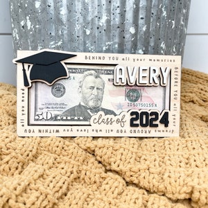 May include: A wooden frame with a graduation cap, the name Avery, and the text "class of 2024" is displayed with a $50 bill inside. The frame is on a tan knitted surface.