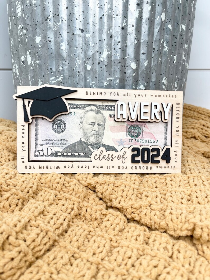 May include: A wooden frame with a graduation cap, the name Avery, and the text "class of 2024" is displayed with a $50 bill inside. The frame is on a tan knitted surface.