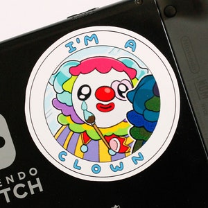 May include: A round sticker featuring a cartoon clown with a red nose, crying a blue tear, and holding a paintbrush. The clown has colorful hair and a purple, yellow, and blue outfit. The text "I'M A CLOWN" is written around the clown.