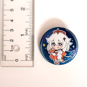 May include: A circular pinback button featuring a stylized white-haired character with orange accents on a dark blue background with yellow stars. A clear plastic ruler is positioned to the left, showing the button has a diameter of 1.5 inches.