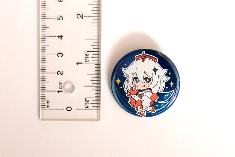 May include: A circular pinback button featuring a stylized white-haired character with orange accents on a dark blue background with yellow stars. A clear plastic ruler is positioned to the left, showing the button has a diameter of 1.5 inches.