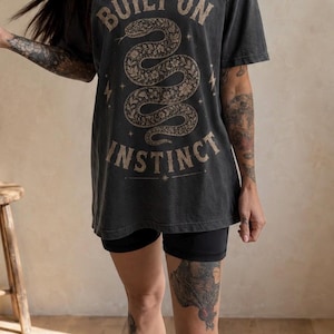 Könnte beinhalten: Ein anthrazitfarbenes Kurzarm-T-Shirt mit einer beigen Grafik einer gewundenen Schlange, umgeben von Blumenmustern und kleinen Sternen. Der Text "BUILT ON INSTINCT" ist in einer fetten, geschwungenen Schrift um die Schlange gedruckt.