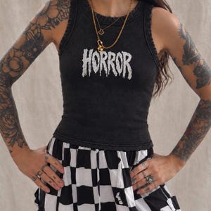 Puede incluir: Una camiseta sin mangas de color gris carbón con lavado ácido que presenta la palabra HORROR impresa en una fuente blanca, desgastada y con efecto de goteo en el pecho. La prenda tiene un cuello redondo acanalado y un corte ajustado, combinada con una falda a cuadros.