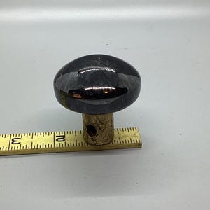 May include: A black ceramic knob with a rounded top and a small hole in the center. The knob is mounted on a wooden base.