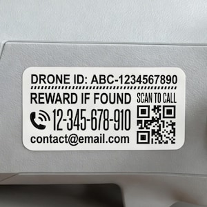 Puede incluir: Una pegatina blanca en un dron con el texto "DRONE ID: ABC-1234567890", "REWARD IF FOUND SCAN TO CALL", un número de teléfono y una dirección de correo electrónico. También hay dos códigos QR.
