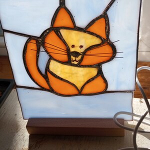 Peut inclure: Une fenêtre en vitrail représentant un chat de dessin animé en verre orange et jaune. Le chat est assis sur un fond blanc et a des moustaches et des yeux noirs.