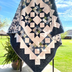 May include: A handmade quilt with a starburst design. The quilt features a white center with navy blue and green accents. The border is navy blue with a small white floral pattern. The quilt is displayed outdoors.