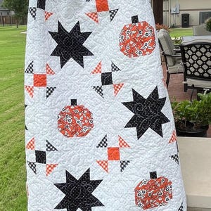 May include: White quilt with a repeating pattern of black stars, orange pumpkins, and orange and black squares. The quilt has a scalloped edge with a black and white checkered pattern. The quilt is hanging outside.