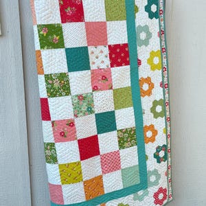 May include: A colorful patchwork quilt with a floral design. The quilt features squares of various colors and patterns, including red, pink, green, and white. The edges are trimmed with a teal border and flower-shaped patterns. The quilt is draped over a wooden stand.