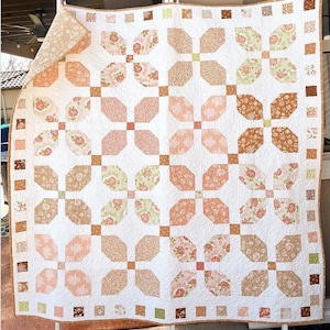 May include: A square quilt featuring a repeating pattern of four-petaled floral blocks in shades of peach, sage green, and tan on a white background. The border consists of small alternating squares in matching colors.