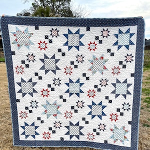 May include: A white quilt with a repeating star pattern in navy blue, red, and light blue. The stars are of varying sizes and patterns, including polka dots and floral prints. The quilt has a navy blue border.