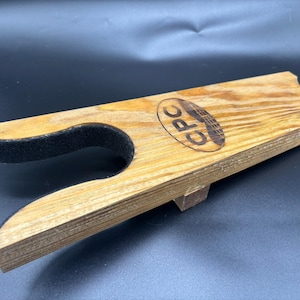 Könnte beinhalten: Stiefelknecht aus Holz mit einem mit schwarzem Filz gefütterten Fußteil. Der Stiefelentferner hat eine natürliche Holzmaserung und die Buchstaben "CPC" sind auf der Oberfläche eingebrannt. Ein kleiner Holzträger ist unten angebracht.