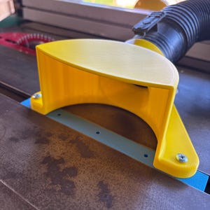 May include: A bright yellow table saw blade guard with a curved design. The guard is attached to the saw with screws and sits over the blade. A black hose is connected to the guard.