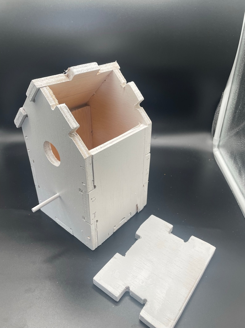 Puede incluir: Casa de p&aacute;jaros de madera blanca con un techo extra&iacute;ble y una peque&ntilde;a percha. La casa de p&aacute;jaros est&aacute; hecha de madera contrachapada y tiene un dise&ntilde;o simple.
