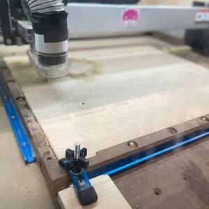 Puede incluir: Primer plano de una m&aacute;quina CNC cortando una pieza de madera. La m&aacute;quina tiene una pista azul y negra y un tubo de pl&aacute;stico transparente conectado a la cabeza de corte. La madera es de color marr&oacute;n claro y tiene algunos agujeros perforados.