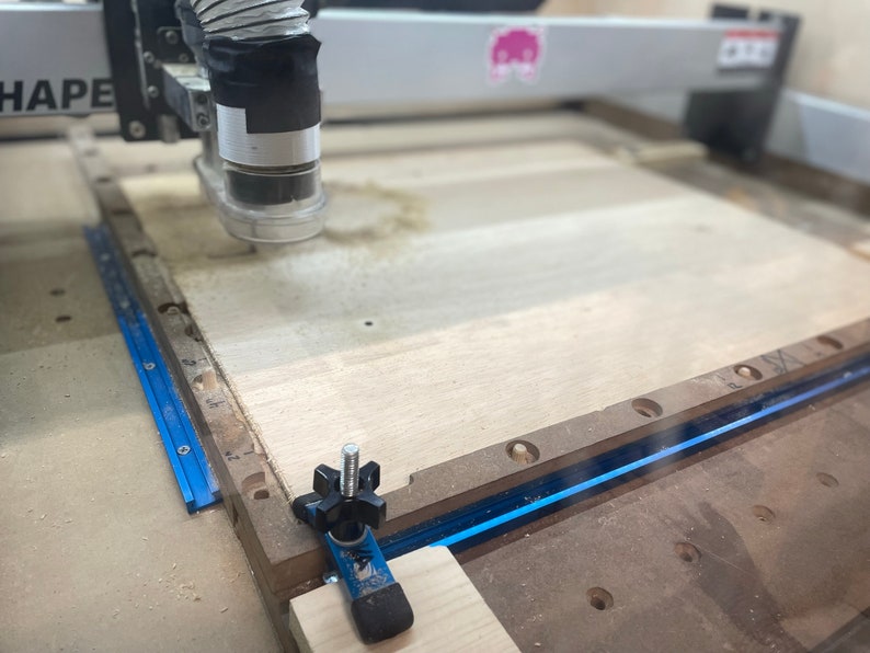 Puede incluir: Primer plano de una m&aacute;quina CNC cortando una pieza de madera. La m&aacute;quina tiene una pista azul y negra y un tubo de pl&aacute;stico transparente conectado a la cabeza de corte. La madera es de color marr&oacute;n claro y tiene algunos agujeros perforados.