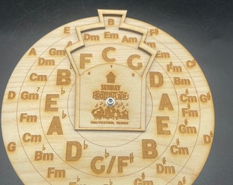 Puede incluir: Una herramienta circular de teoría musical de madera con notaciones musicales y armaduras grabadas. El centro presenta un emblema rectangular con texto y una pequeña perilla de metal. El texto dice "SUNDAY HOOFNANNY GALVESTON, TEXAS."