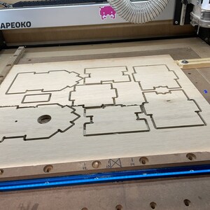 Puede incluir: Primer plano de una tabla de madera con formas recortadas, probablemente para un proyecto de carpinter&iacute;a. La tabla est&aacute; en una m&aacute;quina con una tira de luz azul y el texto "SHAPEOKO" visible.