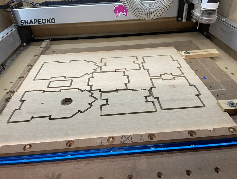 Puede incluir: Primer plano de una tabla de madera con formas recortadas, probablemente para un proyecto de carpinter&iacute;a. La tabla est&aacute; en una m&aacute;quina con una tira de luz azul y el texto "SHAPEOKO" visible.