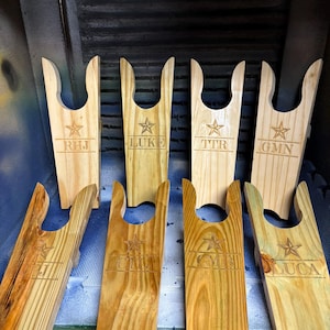 Könnte beinhalten: Holz-Stiefelknechte in verschiedenen Brauntönen. Jeder Stiefelknecht hat ein Sterndesign und Initialen oder Namen, darunter "RHJ", "LUKE", "TTR", "GMN", "CJM" und "LUCA". Die Stiefelknechte sind auf einer blauen Oberfläche angeordnet.