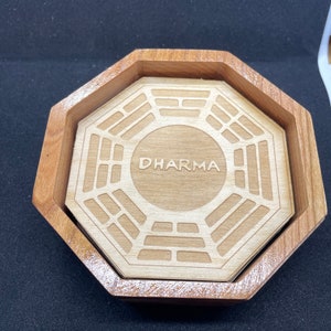Pode incluir: Um descanso de copo de madeira com o logotipo da Iniciativa Dharma e a palavra "DHARMA" gravada no centro.